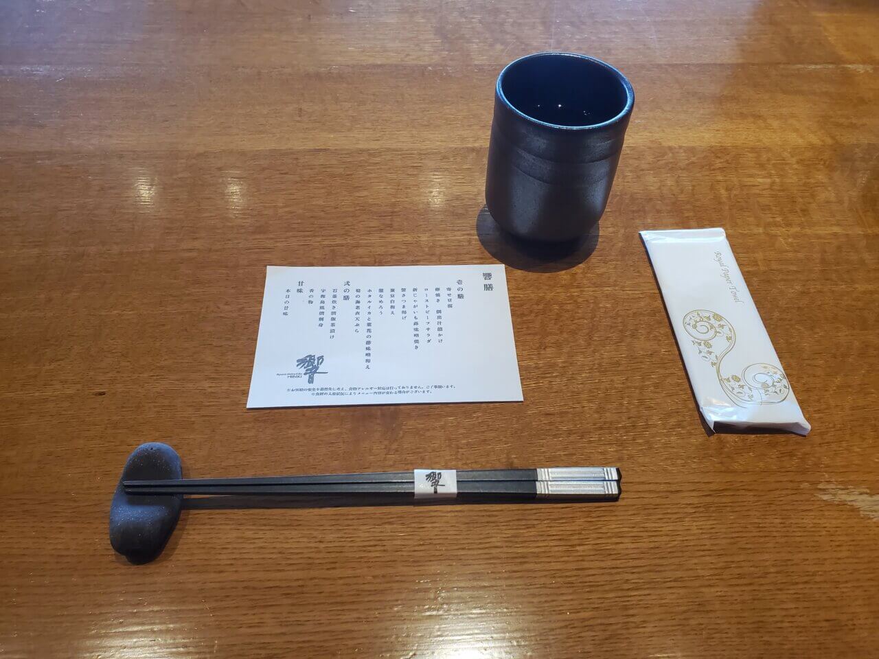 卓上に整えられた箸、箸置き、湯飲み、おしぼり、そしてメニューカードが置かれている様子。木目のテーブルがきれいに磨かれている。